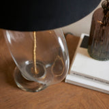 Close-up of the Endon Bramleigh table lamp's clear glass ellipsoid base and black shade on a wood surface beside stacked books and a brown glass vessel.