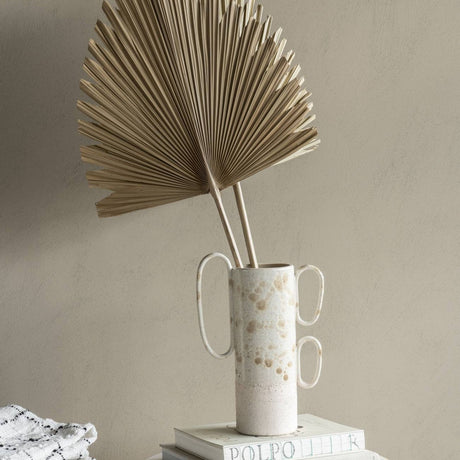 Libra Interiors Arrowhead Fan Palm Leaf artificial stems arranged in a speckled ceramic vase with looped handles, set on stacked books against a neutral wall.