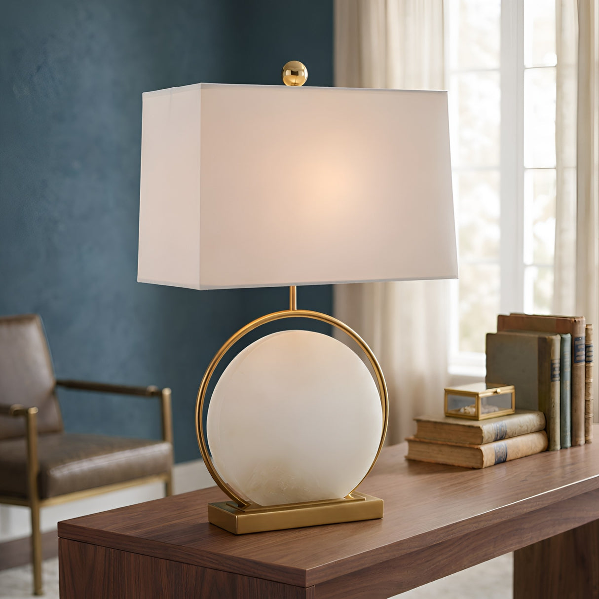 The Orivane 1 Light Circular Alabaster Table Lamp in Brass & Soft White sits on a wooden desk next to stacked books, with a chair and window in the background. Its sculptural brass base and soft white shade add modern elegance to the space.
