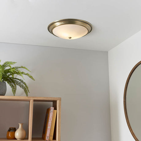 Atvorne Flush Ceiling Light with beaded antique brass surround and frosted glass diffuser, shown lit on a white ceiling above a hallway shelf and mirror.