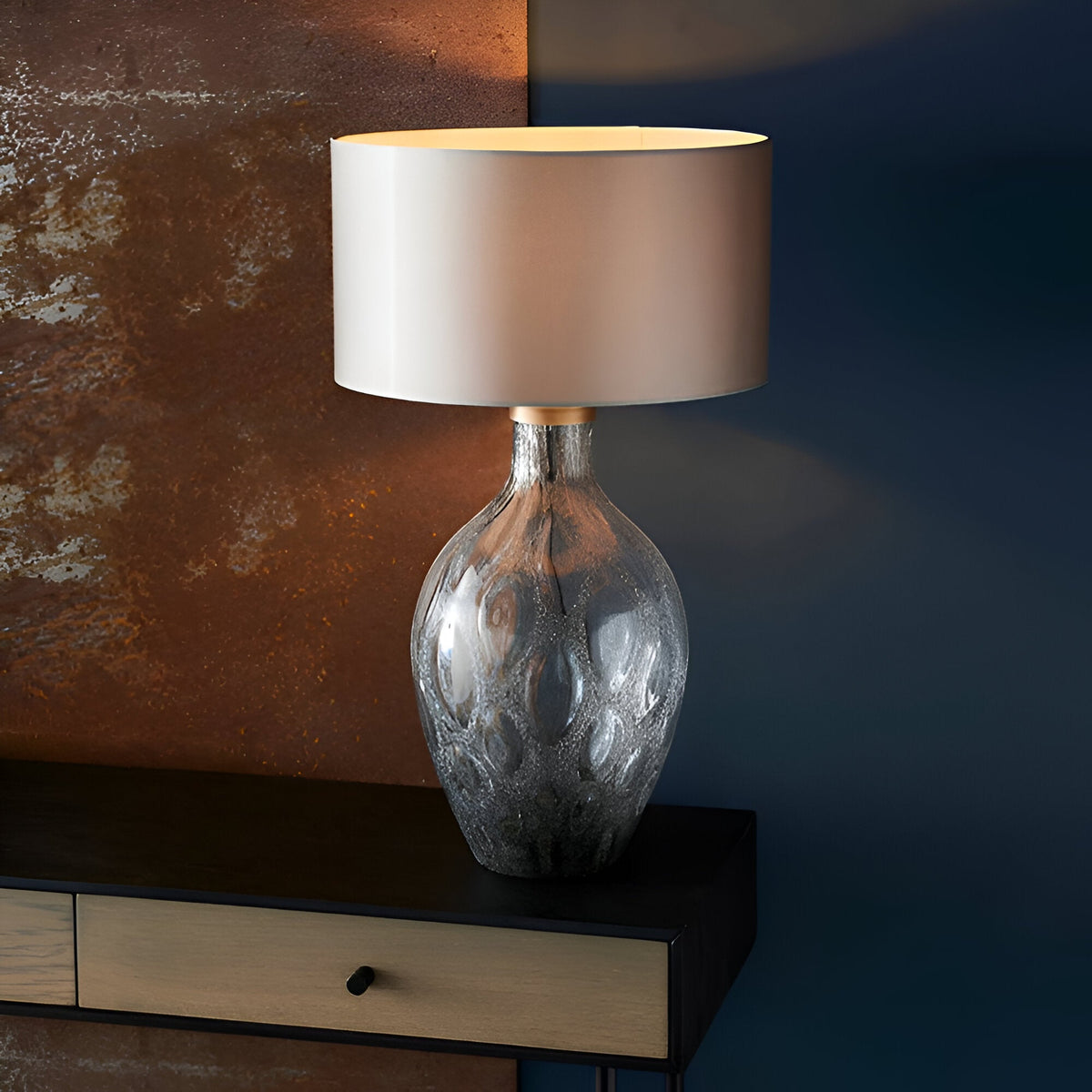 Endon Brenstead table lamp with charcoal-tinted textured artisan glass base, antique brass detail and cream drum shade, lit on a dark console.