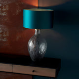 Endon Brenstead table lamp with textured charcoal artisan glass base and antique brass detail, topped with a teal satin drum shade on a dark console.