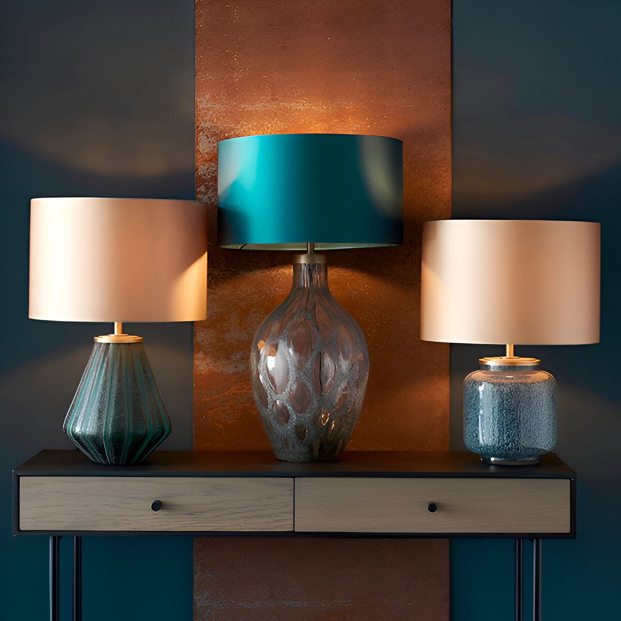 Endon Brenstead trio of table lamps with textured glass bases in charcoal and teal tones, topped with drum shades, on a wooden console against a copper panel.