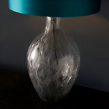 Endon Brenstead table lamp with textured charcoal-tinted artisan glass base and teal satin drum shade on a dark wood surface.