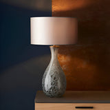 Carldale Table Lamp in Bronze with grey cloud-effect glass hourglass base, brushed metalwork, and pale drum shade, against a dark blue and copper wall.