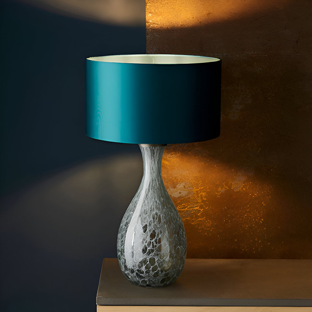 Carldale Table Lamp in Bronze with grey cloud-effect glass hourglass base, paired with a teal drum shade, styled against a dark and amber-lit wall.