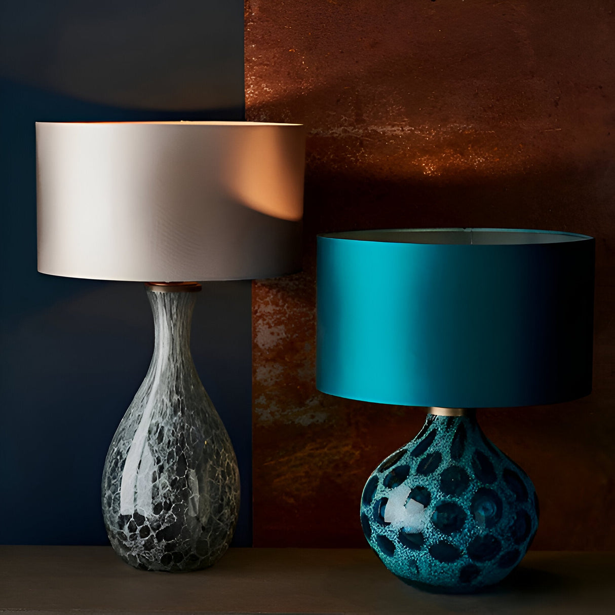 Carldale Table Lamp in bronze with grey cloud-effect glass hourglass base, shown left with a taupe drum shade beside a teal lamp on a dark background.