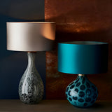 Carldale Table Lamp in bronze with grey cloud-effect glass hourglass base, shown left with a taupe drum shade beside a teal lamp on a dark background.