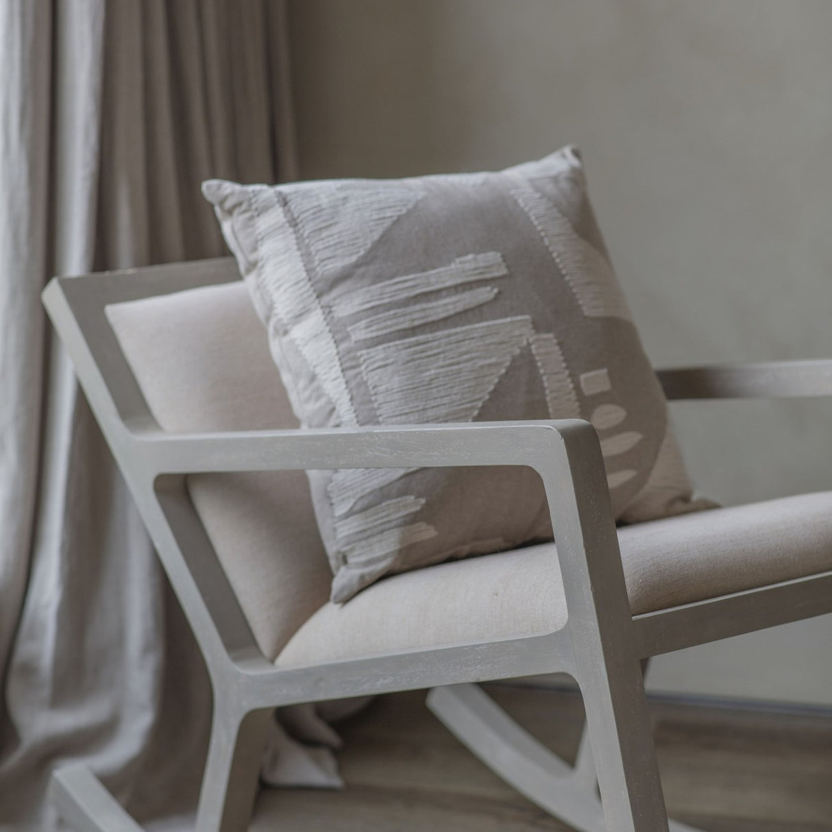 Libra Interiors textured cotton 50cm square cushion in beige with cream stitched abstract motif, resting on a pale wooden rocking chair by linen curtains.