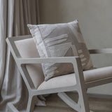 Libra Interiors textured cotton 50cm square cushion in beige with cream stitched abstract motif, resting on a pale wooden rocking chair by linen curtains.