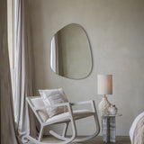 Libra Interiors Textured Cotton 50cm square cushion in natural beige with cream stitched motif, on a white rocking chair beside a shaped wall mirror and lamp.