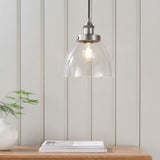 Wentworth Pendant Light in silver with clear dome glass shade and exposed filament bulb, hung above a wooden table in a room with panelled walls.