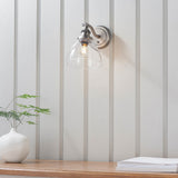 Silver Wentworth wall light with clear glass dome shade and adjustable knuckle arm, shown lit above a wooden shelf with a vase and plant.
