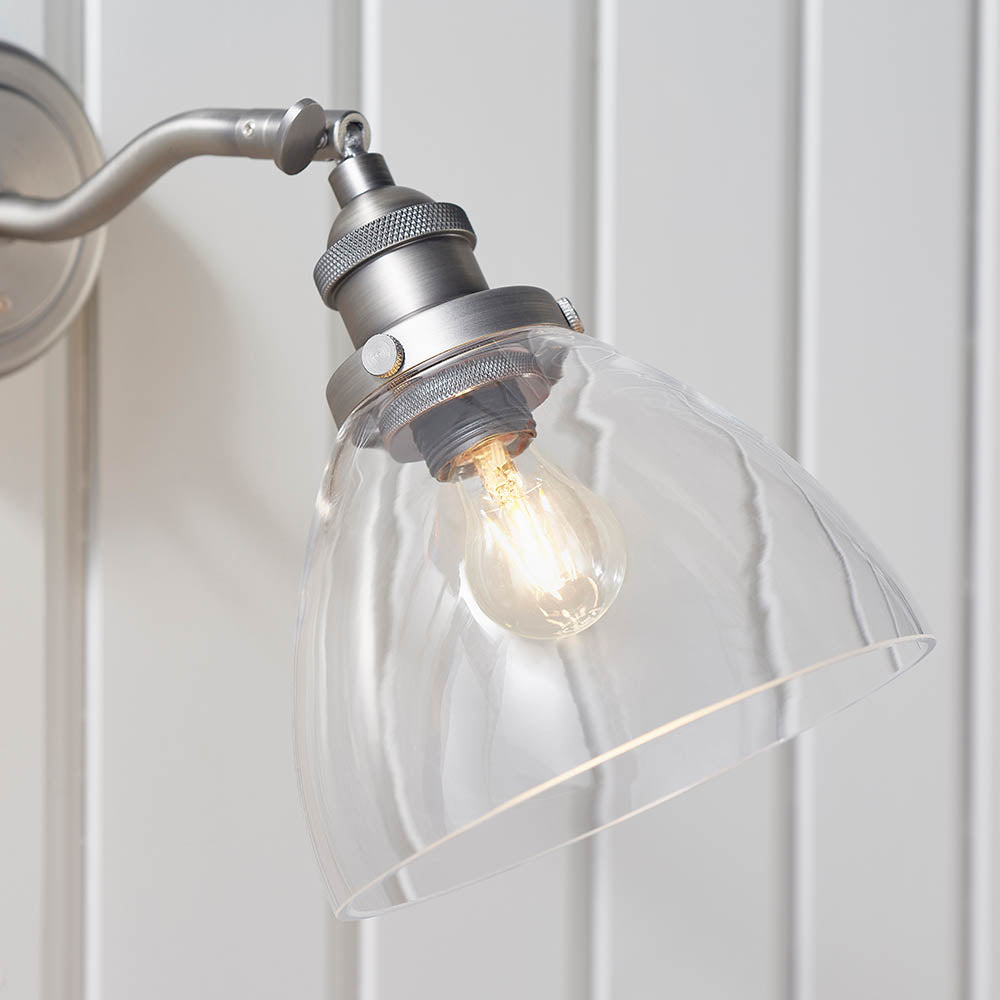 Wentworth Wall Light in silver with adjustable knuckle joint, knurled lamp holder, and clear glass dome shade, shown lit against a white panelled wall.