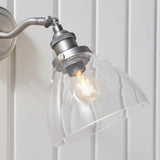 Wentworth Wall Light in silver with adjustable knuckle joint, knurled lamp holder, and clear glass dome shade, shown lit against a white panelled wall.