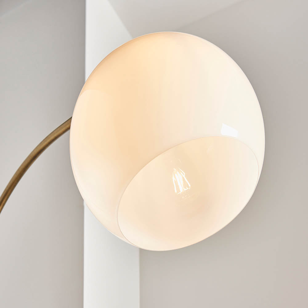 Close-up of Sinclair Floor Lamp's round gloss white glass shade with concave center revealing bulb, on an antique brass curved arm.