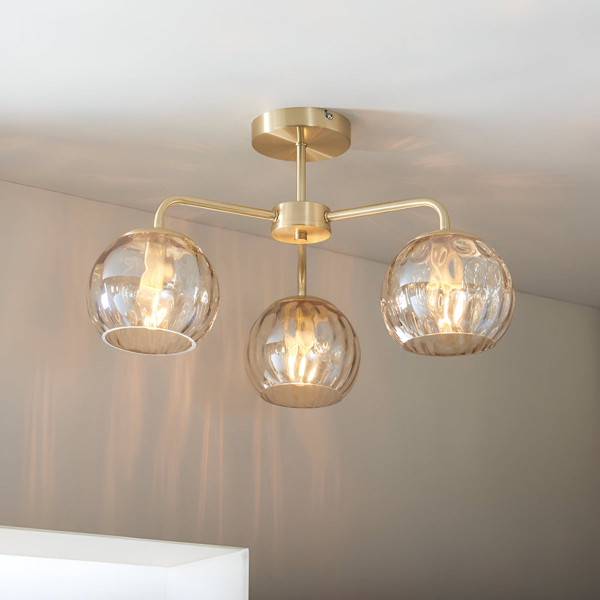 Endon Everly 3 light semi-flush ceiling light in gold, with three arms holding dimpled amber glass globe shades beneath a circular brass plate.