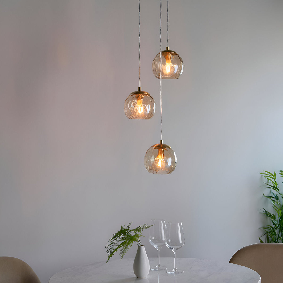 Draycott 3 Light Dimpled Glass Pendant in Satin Brass, three round amber glass shades at staggered heights, shown above a dining table.