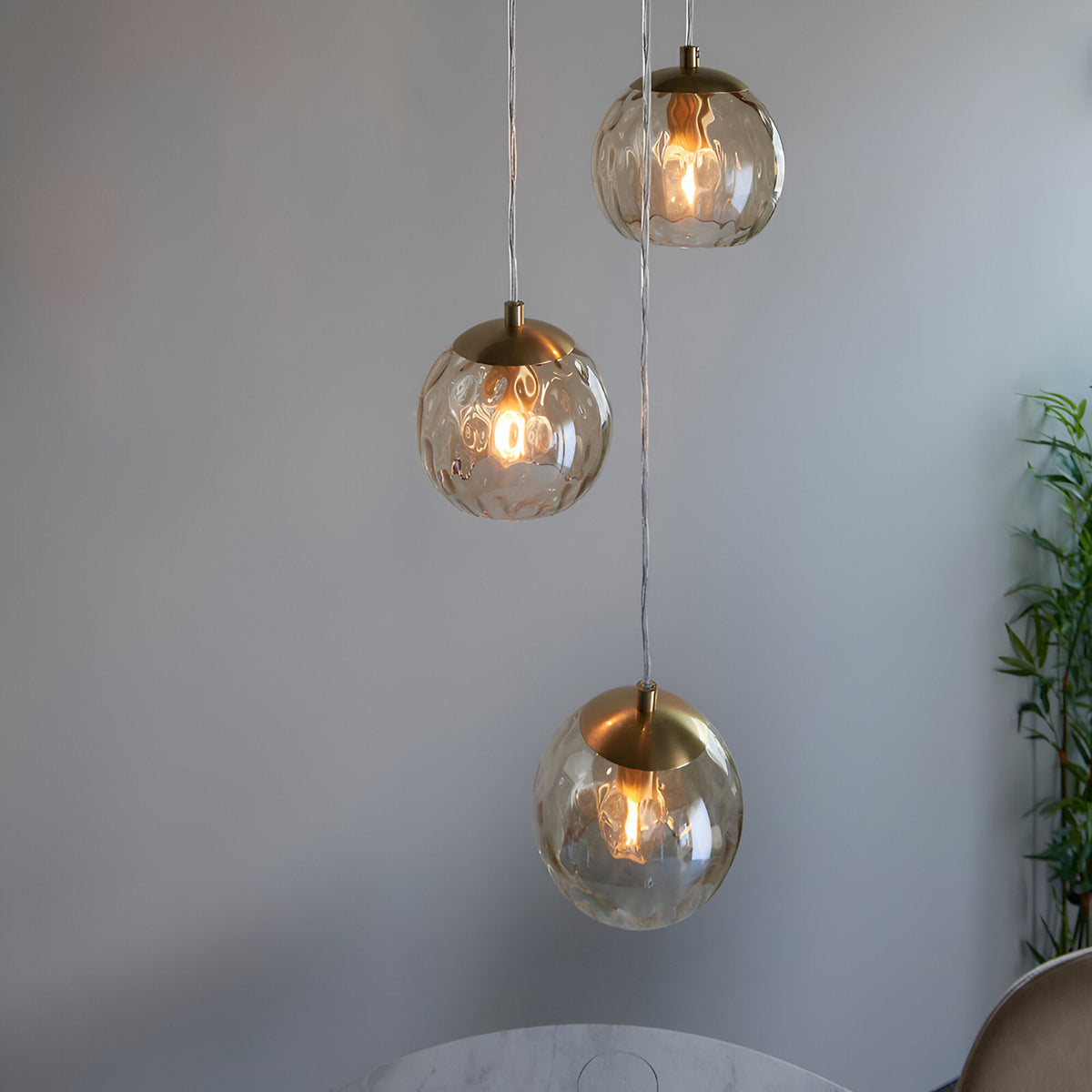 Draycott 3 Light Dimpled Glass Pendant in satin brass, three round champagne glass globes at staggered heights on clear cables, shown against a plain grey wall.