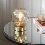 Draycott Dimpled Glass Table Lamp in Satin Gold with champagne-tinted globe shade, lit on a glass side table beside books and a bowl.