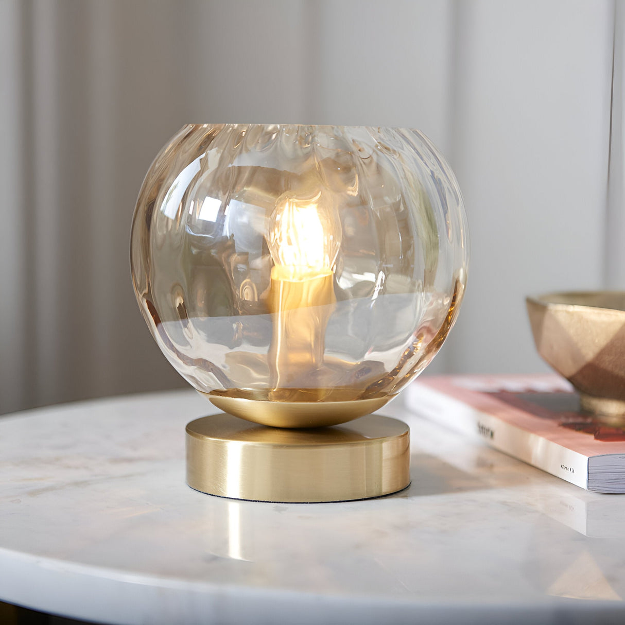 Draycott Dimpled Glass Table Lamp in Satin Gold, lit on a marble side table with a satin gold base and champagne-toned globe shade beside books.