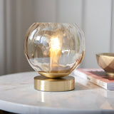 Draycott Dimpled Glass Table Lamp in Satin Gold, lit on a marble side table with a satin gold base and champagne-toned globe shade beside books.