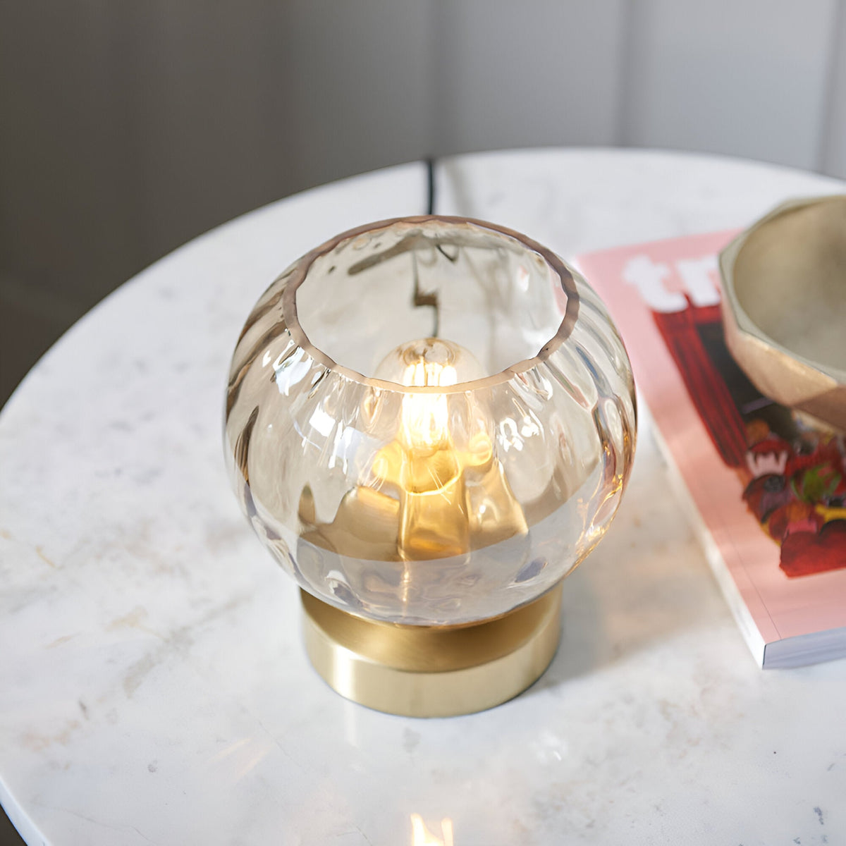 Draycott Dimpled Glass Table Lamp in champagne glass with satin gold base, lit on a marble side table beside a magazine and bowl.