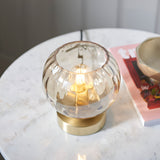 Draycott Dimpled Glass Table Lamp in champagne glass with satin gold base, lit on a marble side table beside a magazine and bowl.