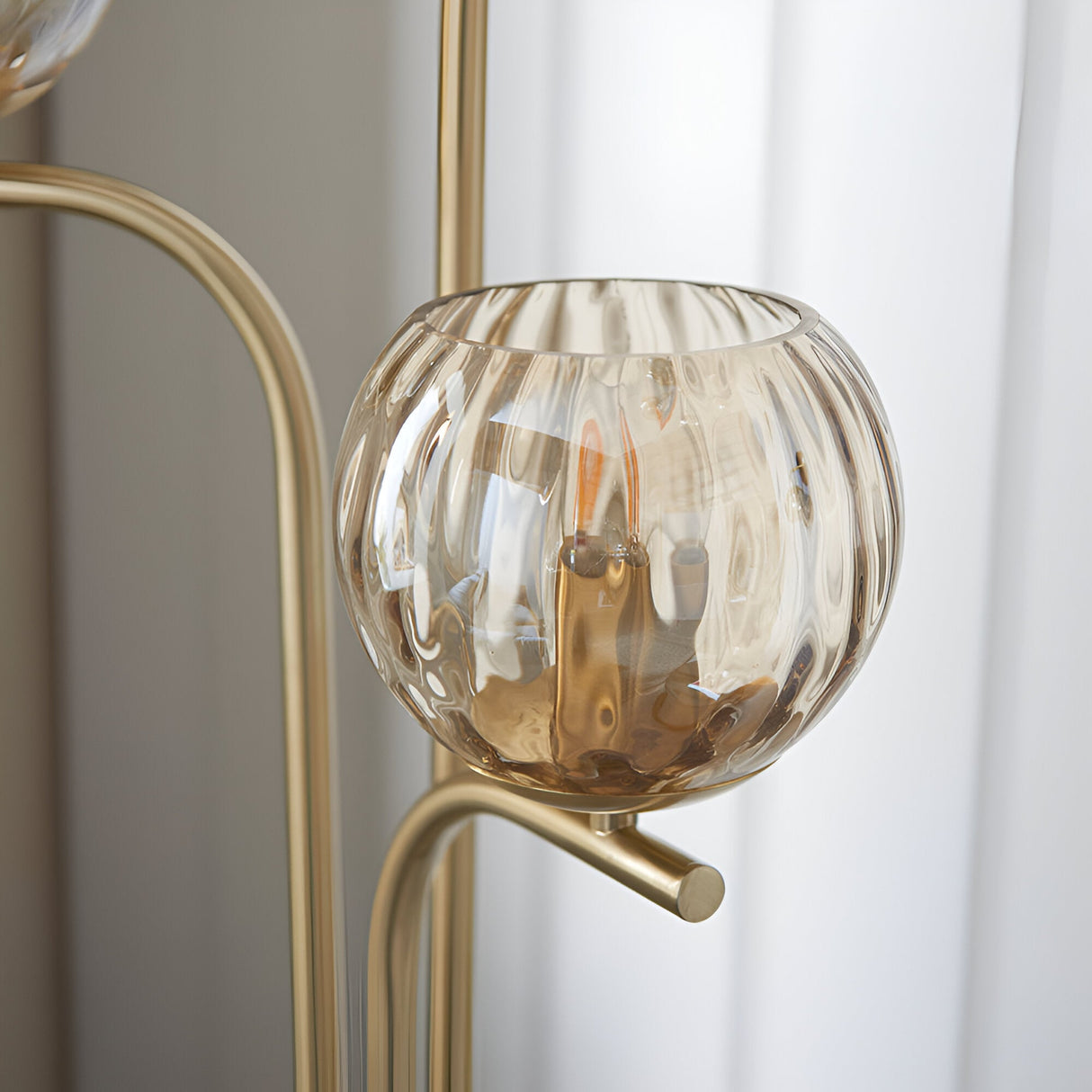 Close-up of the Endon Draycott floor lamp shade, a champagne dimpled glass globe cradled on a curved satin brass arm beside sheer curtains.