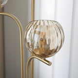 Close-up of the Endon Draycott floor lamp shade, a champagne dimpled glass globe cradled on a curved satin brass arm beside sheer curtains.