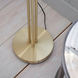 Close-up of the Endon Draycott floor lamp's round satin brass base and three-rod stem on pale wooden flooring, with black flex trailing behind.