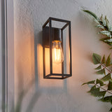 Brenford Outdoor Wall Light in black, an open rectangular aluminium cage design with visible E27 filament bulb, mounted on a textured wall near foliage.