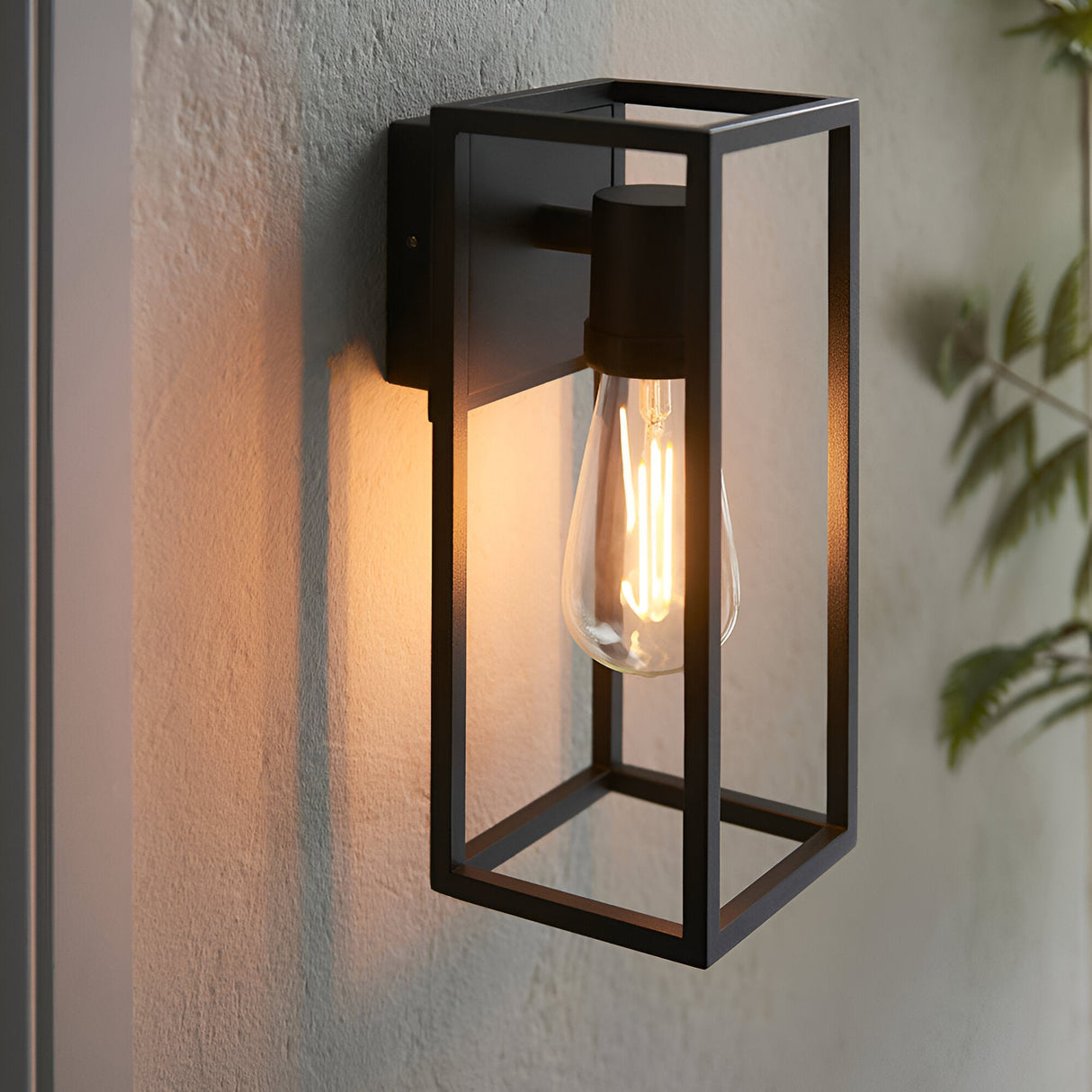 Brenford Outdoor Wall Light in black, an open-frame lantern-style aluminium fitting with visible filament bulb, mounted on a textured wall.