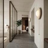 A modern minimalist hallway features light gray walls, large floor tiles, a potted plant, the Solenne 1 Light Travertine Wall Light With Pull Switch in light beige, and glass-paneled partitions separating it from the living area and bedroom.