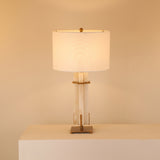 Pilaster table lamp with a natural alabaster column in a brushed brass frame, topped with a white fabric drum shade, glowing softly on a light plinth.
