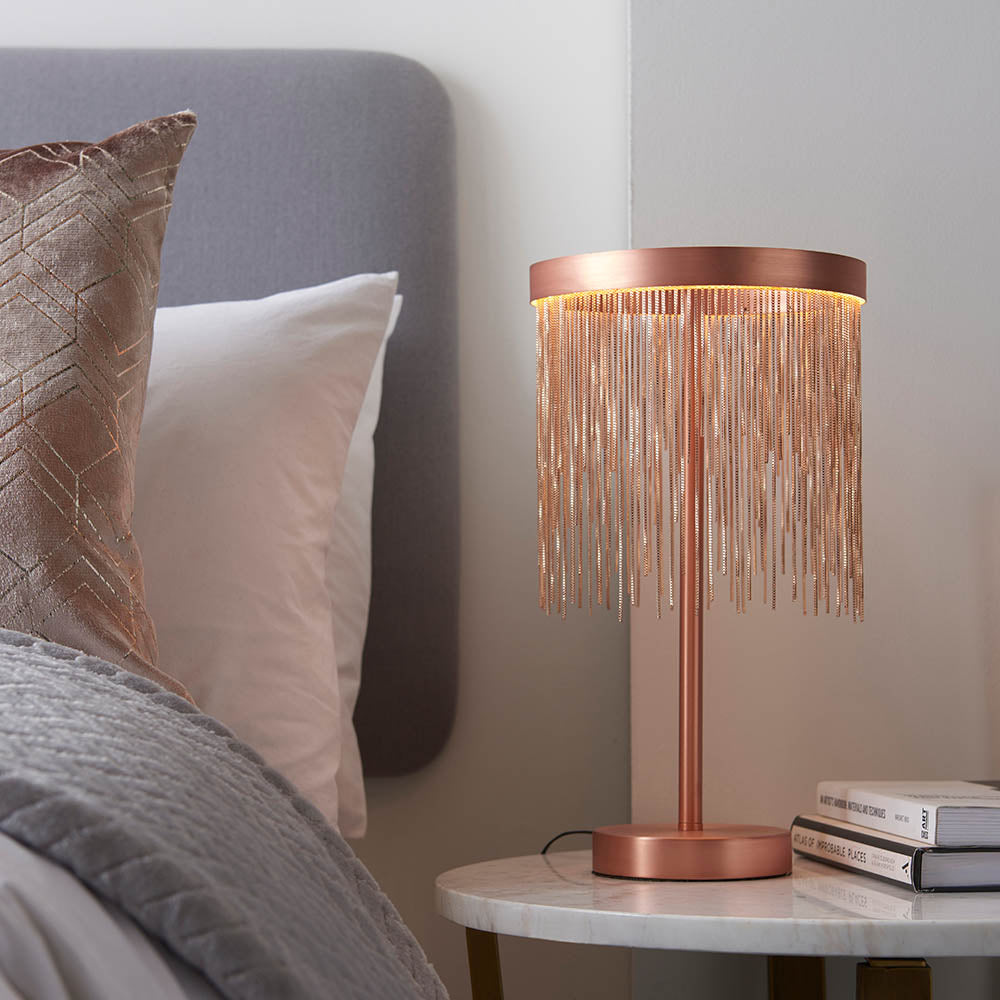 Endon Danestead brushed copper LED table lamp with cascading copper-toned chains on a marble bedside table beside books and a grey upholstered bed.