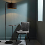 Caldfield Floor Lamp in satin black metal with curved stem, built-in side table, and black fabric shade, shown beside a grey upholstered chair.