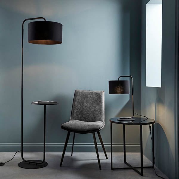 Caldfield Floor Lamp in black metal with a curved arm, black fabric cylinder shade, and built-in round table top, shown beside a chair in a living room setting.