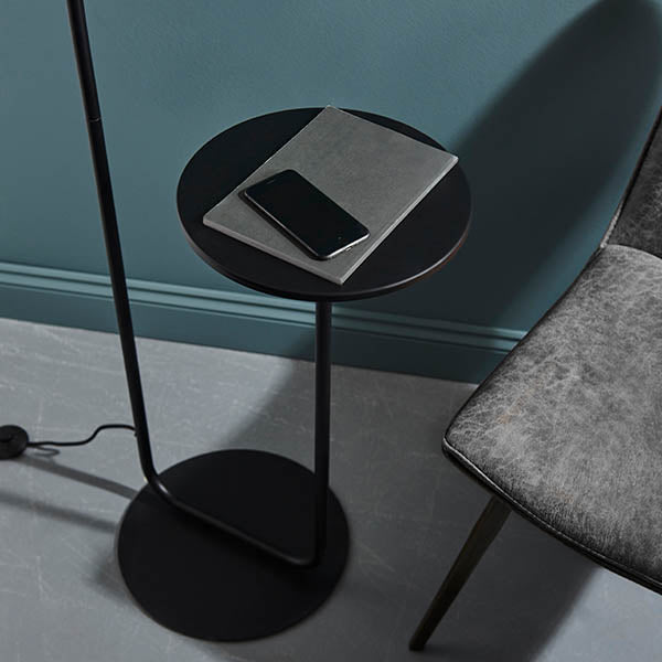 Caldfield Floor Lamp in satin black metal with round base and built-in side table holding a notebook and phone, shown against a teal wall.