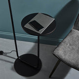 Caldfield Floor Lamp in satin black metal with round base and built-in side table holding a notebook and phone, shown against a teal wall.