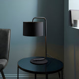 Caldfield Table Lamp in satin black with a curved metal stem and cylindrical fabric shade, shown on a dark round side table in a muted living room setting.