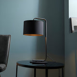 Caldfield Table Lamp in satin black with cylinder fabric shade and angular metal base, styled on a black side table beside a dark chair.