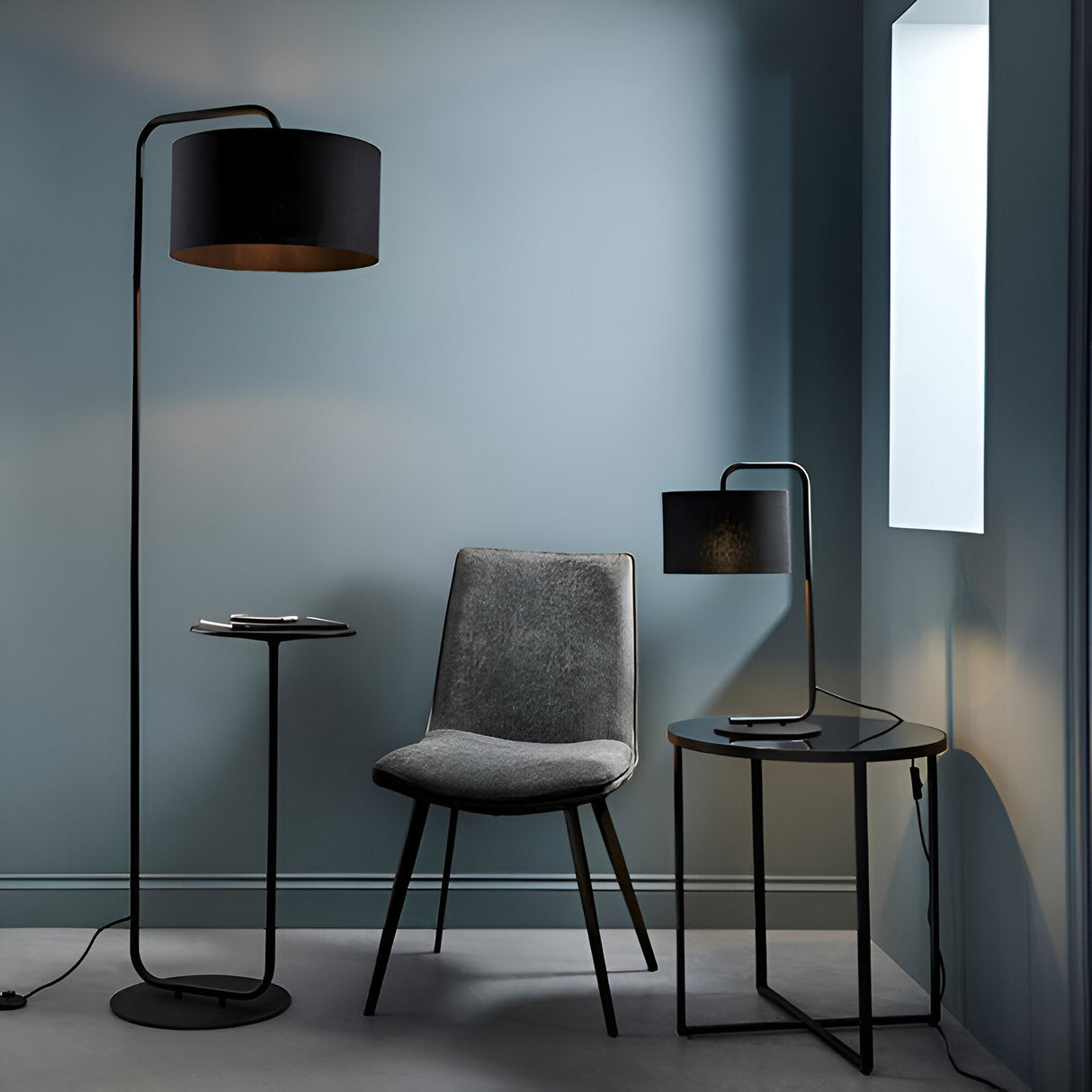 Caldfield Table Lamp in satin black metal with cylinder fabric shade, shown on a black side table beside a grey chair in a living room setting.