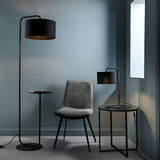 Caldfield Table Lamp in satin black metal with cylinder fabric shade, shown on a black side table beside a grey chair in a living room setting.