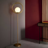 Endon Brackshaw floor lamp with opal glass orb inside three brushed gold rings on a slim stem, styled in a corner beside a dark red wall.