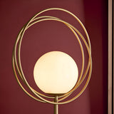 Endon Brackshaw floor lamp close-up showing an opal glass orb held within three interlinking brushed gold rings against a deep burgundy wall.