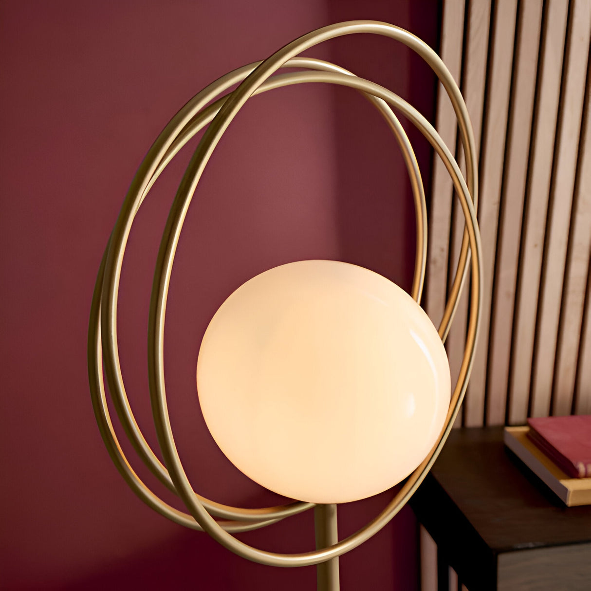 Endon Brackshaw floor lamp with opal glass orb held in three interlinking brushed gold rings, shown lit against a burgundy wall and wood slat panel.