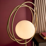 Endon Brackshaw floor lamp with opal glass orb held in three interlinking brushed gold rings, shown lit against a burgundy wall and wood slat panel.