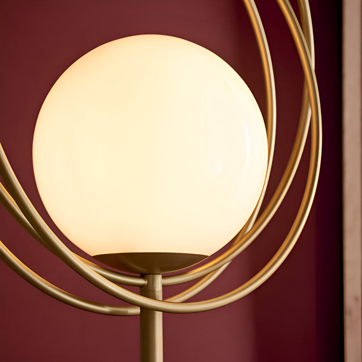 Close-up of the Endon Brackshaw floor lamp's opal glass orb held within three interlinking brushed gold rings against a deep burgundy wall.