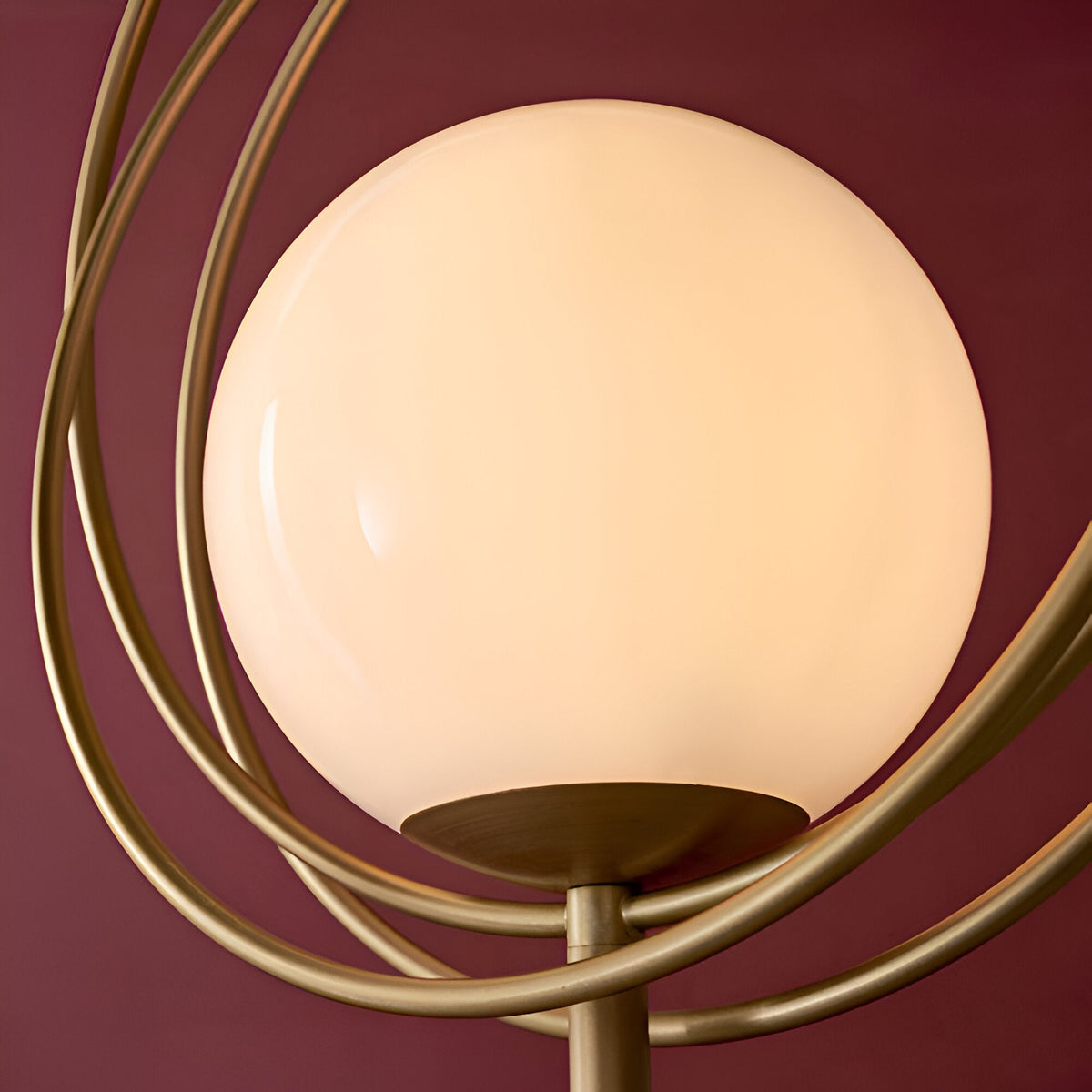 Close-up of the Endon Brackshaw floor lamp showing its opal glass orb cradled within three interlinking brushed gold rings against a deep burgundy wall.